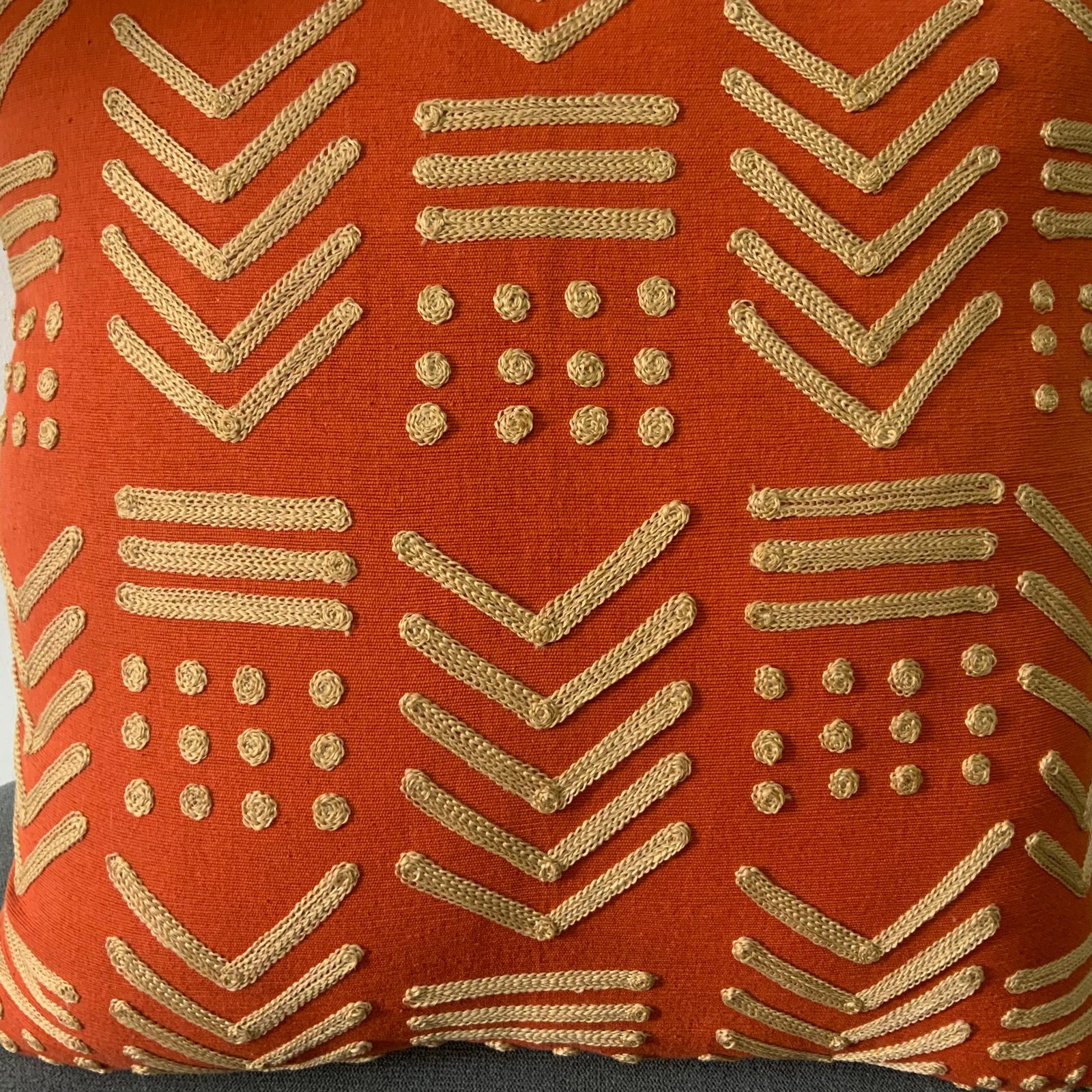 Square terracotta orange throw pillow  with raised gold-toned geometric embroidery in a tribal mudcloth design featuring chevrons and dots.